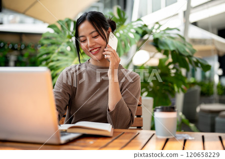 A beautiful Asian woman talking on the phone while working remotely from a cafe. A beautiful Asian woman talking on the phone while working remotely from a cafe. 120658229