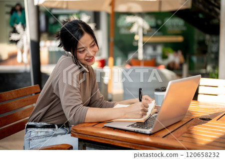 A beautiful Asian woman is taking notes in a book while talking on the phone, working remotely. A beautiful Asian woman is taking notes in a book while talking on the phone, working remotely. 120658232