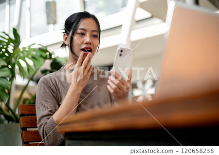 An amazed, excited Asian woman surprised by an unexpected good news through a message on her phone. An amazed, excited Asian woman surprised by an unexpected good news through a message on her phone. 120658238