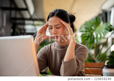 A stressed Asian woman sits at an outdoor table, rubs her temples, feeling a headache while working. 120658246