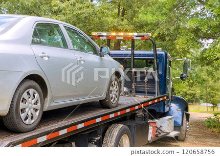 Silver sedan is being loaded onto tow truck in wooded area during truck stands ready for transport 120658756