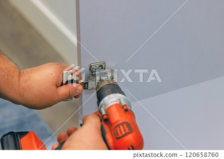Worker uses an electric drill to securely attach kitchen door hinge to wooden contemporary indoor setting 120658760