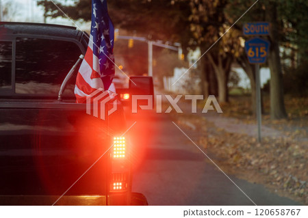 Pickup truck displaying an American flag travels along calm road surrounded by trees warm glow of dusk enhances peaceful atmosphere of autumn evening. 120658767