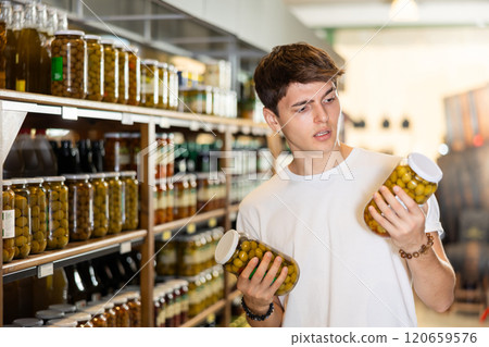 Interested guy choosing pickled olives in glass jars in supermarket Interested guy choosing pickled olives in glass jars in supermarket 120659576