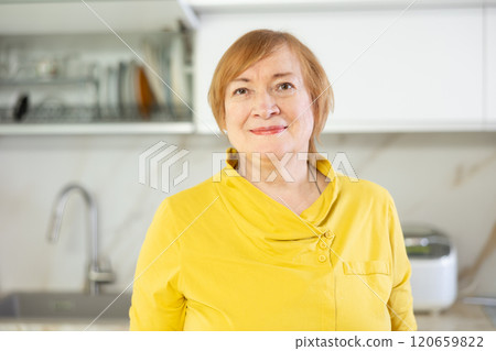 Smiling senior woman in casual clothes standing in home kitchen with open arms 120659822