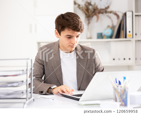 Young man office manager sitting at table and using laptop computer Young man office manager sitting at table and using laptop computer 120659958