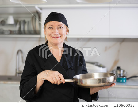 Happy mature female cook in uniform standing and showing her kitchen Happy mature female cook in uniform standing and showing her kitchen 120660033