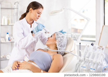 Cosmetologist applying cosmetic products on facial skin of female patient with airbrush 120660100