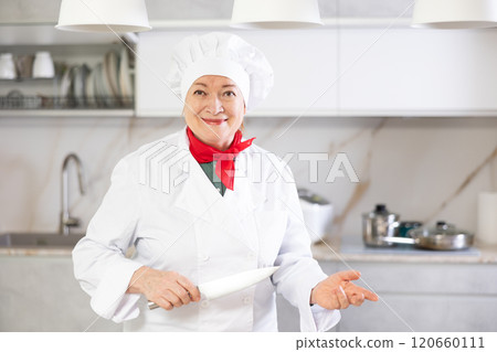 Cheerful mature female chef standing in kitchen ready to give cooking lesson 120660111