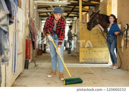 Positive young girl horse farm worker cleaning floor at stable. Adult woman holds horse by reins in background 120660232