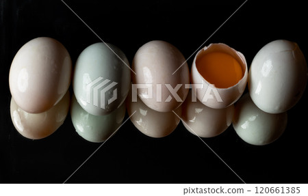 Five raw duck eggs are sitting in a row with an egg broken in half showing the yolk and white egg with reflection on dark background. Five raw duck eggs are sitting in a row with an egg broken in half showing the yolk and white egg with reflection on dark background. 120661385