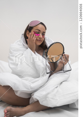 Cocooned in her blanket, a woman applies eye patches while seated on her bedroom bed, meticulously inspecting her face with a hand mirror. Cocooned in her blanket, a woman applies eye patches while seated on her bedroom bed, meticulously inspecting her face with a hand mirror. 120664008
