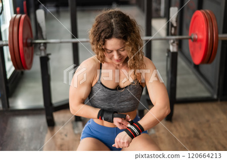 Caucasian forty-year-old woman puts on wrist straps before doing barbell exercises in the gym.  120664213