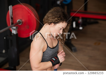 Caucasian forty-year-old woman puts on wrist straps before doing barbell exercises in the gym. Caucasian forty-year-old woman puts on wrist straps before doing barbell exercises in the gym. 120665233