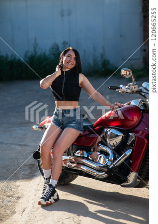 Brunette woman in shorts posing on a red motorcycle. Vertical photo. Brunette woman in shorts posing on a red motorcycle. Vertical photo. 120665255
