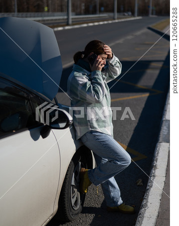 Sad Caucasian woman calling on phone while standing near car with open hood. Sad Caucasian woman calling on phone while standing near car with open hood. 120665288