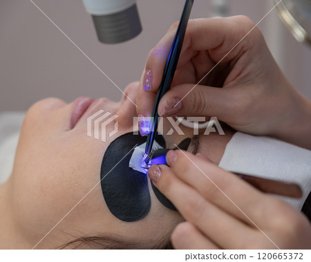 A woman undergoing eyelash extension procedure using an ultraviolet lamp. A woman undergoing eyelash extension procedure using an ultraviolet lamp. 120665372