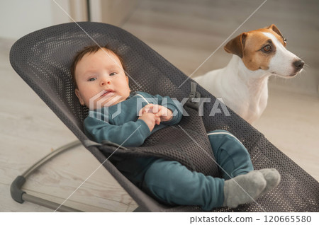 A dog sits next to a cute three-month-old boy dressed in a blue overalls in a baby lounger. 120665580