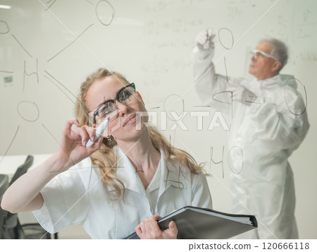 A woman chemist writes a formula on glass. An elderly Caucasian man in a protective suit is doing tests. 120666118