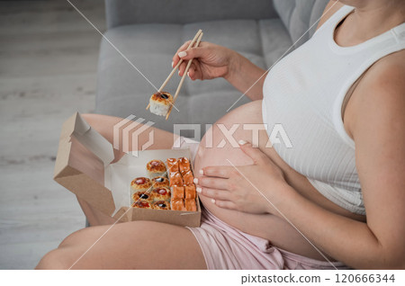 A pregnant woman sits on the sofa and eats rolls from a box. Food delivery. Close up of belly. A pregnant woman sits on the sofa and eats rolls from a box. Food delivery. Close up of belly. 120666344