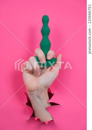 Female hand sticking out with green anal beads from pink paper background. Vertical photo. Female hand sticking out with green anal beads from pink paper background. Vertical photo. 120666761