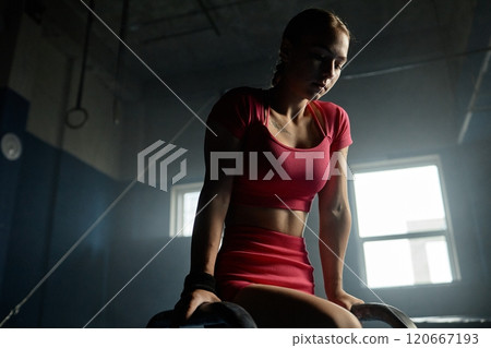 Medium shot of low spirited athletic woman in seated position on floor bars thinking about challenging competition in gymnastics while working out in hard light casting through gym window, copy space Medium shot of low spirited athletic woman in seated position on floor bars thinking about challenging competition in gymnastics while working out in hard light casting through gym window, copy space 120667193