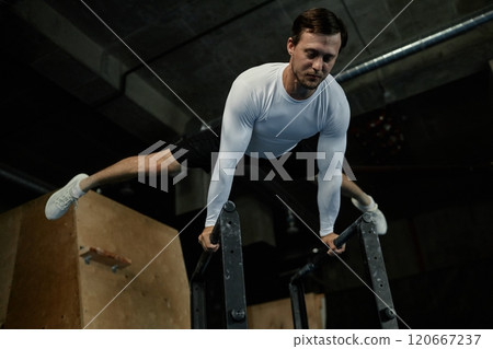 Low angle shot of professional male gymnast in white long sleeved shirt perfectly holding straddle planche on parallel bars while performing calisthenics in gym with low key lighting, copy space 120667237