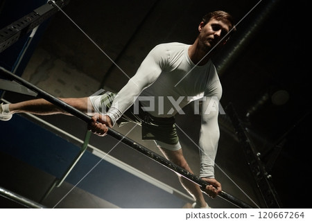 Low angle shot of professional male gymnast performing straddle planche on high bar holding body in static position while exercising calisthenics in gym with low key lighting, copy space 120667264