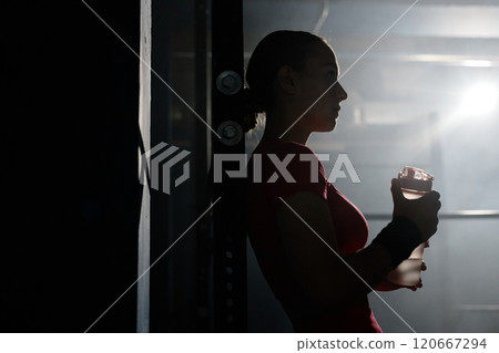 Silhouette of young woman standing against backlight holding bottle of sport drink while having break after intense workout in gym with low key lightning, copy space 120667294