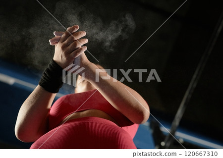 Low angle shot of unrecognizable female gymnast applying magnesia powder on palms clapping hands in chalk cloud in low key lit gym, copy space Low angle shot of unrecognizable female gymnast applying magnesia powder on palms clapping hands in chalk cloud in low key lit gym, copy space 120667310