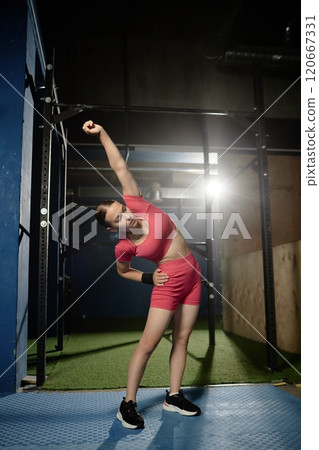 Vertical shot of young sporty woman wearing pink crop top and matching shorts warming up side bending while stretching muscles for daily training on bars in gym, copy space 120667331