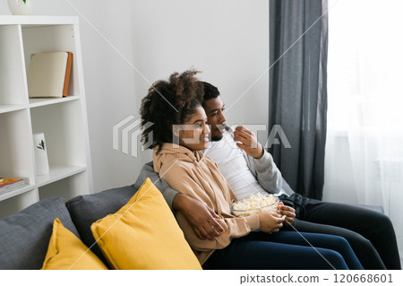 African American couple wife husband watching movie. Family pastime and leisure, watch television, rest. Relaxed man and woman watching TV series together sitting on couch at home enjoying eating African American couple wife husband watching movie. Family pastime and leisure, watch television, rest. Relaxed man and woman watching TV series together sitting on couch at home enjoying eating 120668601