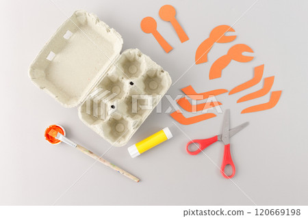 a variety of crafting supplies neatly arranged on a light gray surface. A egg carton, orange paper cutouts shaped, red-handled scissors, a glue stick, a paintbrush, orange paint. DIY project, 120669198