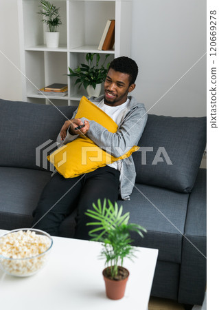 Cheerful curly haired ethnic african american male in casual clothes resting at home on comfortable sofa while watching tv channels or movie during weekend. Tv set, online television and entertiment Cheerful curly haired ethnic african american male in casual clothes resting at home on comfortable sofa while watching tv channels or movie during weekend. Tv set, online television and entertiment 120669278