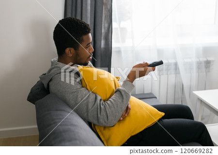 Cheerful curly haired ethnic african american male in casual clothes resting at home on comfortable sofa while watching tv channels or movie during weekend. Tv set, online television and entertiment 120669282