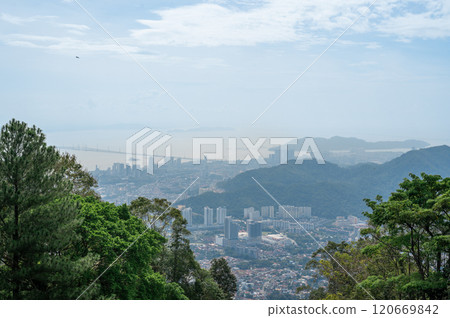 Stunning panoramic view of GeorgeTown Malaysia from hilltop, featuring lush green forest foreground, high-rise buildings, serene sea, cloudy sky, harmonious blend of urban and natural landscapes 120669842