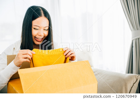Sitting on a couch an Asian woman unboxes a delivered dress from online shopping. Her smiling face exhibits happiness and contentment with the received item. Satisfied buyer concept. Sitting on a couch an Asian woman unboxes a delivered dress from online shopping. Her smiling face exhibits happiness and contentment with the received item. Satisfied buyer concept. 120670949