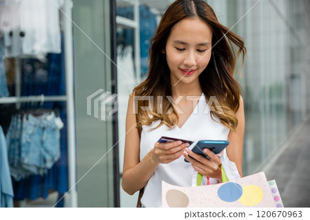 Urban woman enjoying shopping spree with credit card and smartphone in hand. Mall and store windows in the background. Shopping is the therapy she needs. 120670963