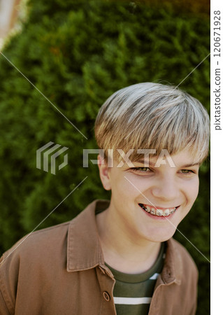 Vertical closeup of joyful teen boy with braces smiling while standing against green plant outdoors Vertical closeup of joyful teen boy with braces smiling while standing against green plant outdoors 120671928