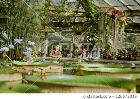 Wide shot of young teacher pointing finger showing yellow flowers to her teen students during tour in botanical garden greenhouse 120672039
