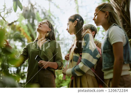 Low angle view of teen students visiting botanical garden, listening to female specialist speaking about plants growing in greenhouse 120672284