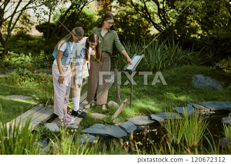 High angle view of young woman working in botanical garden showing aquatic plants to teen girls, copy space High angle view of young woman working in botanical garden showing aquatic plants to teen girls, copy space 120672312