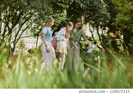 Wide shot of young Caucasian woman working in modern botanical garden showing rare plants to teen schoolgirls Wide shot of young Caucasian woman working in modern botanical garden showing rare plants to teen schoolgirls 120672315