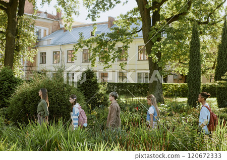 Side view of teenagers following their young teacher, walking along local botanical garden during outdoor lesson 120672333