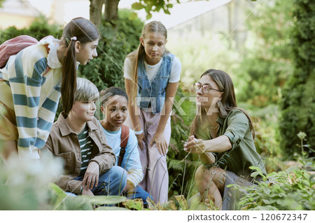 Young female specialist working in local botanical garden showing rare plants to group of teen students 120672347