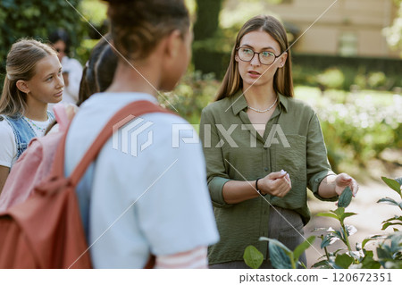 Young female teacher wearing eyeglasses speaking about plants during outdoor class in botanical garden 120672351