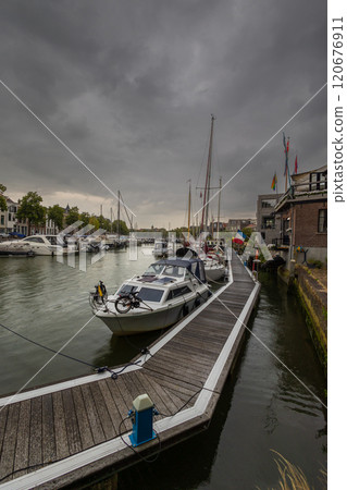 Serene harbor with moored boats under a dramatic cloudy sky. Quaint historic buildings and trees line the waterfront, reflecting in the calm green water, creating peaceful scene. Dordrecht landscape. Serene harbor with moored boats under a dramatic cloudy sky. Quaint historic buildings and trees line the waterfront, reflecting in the calm green water, creating peaceful scene. Dordrecht landscape. 120676911