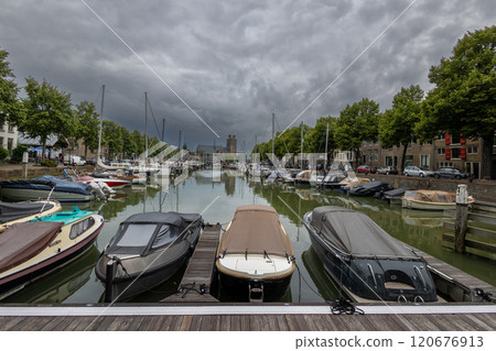 Serene harbor with moored boats under a dramatic cloudy sky. Quaint historic buildings and trees line the waterfront, reflecting in the calm green water, creating peaceful scene. Dordrecht landscape. 120676913