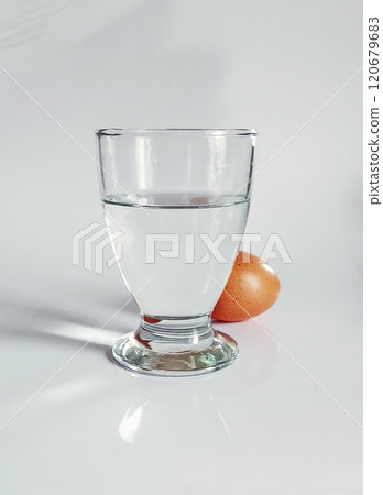 Glass of water casting rays of light on a table and a brown shell egg lying behind it Glass of water casting rays of light on a table and a brown shell egg lying behind it 120679683