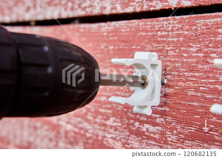 Plumber is using cordless drill to secure white pipe clips to weathered red wooden house for organizing pipeline outdoors. 120682135